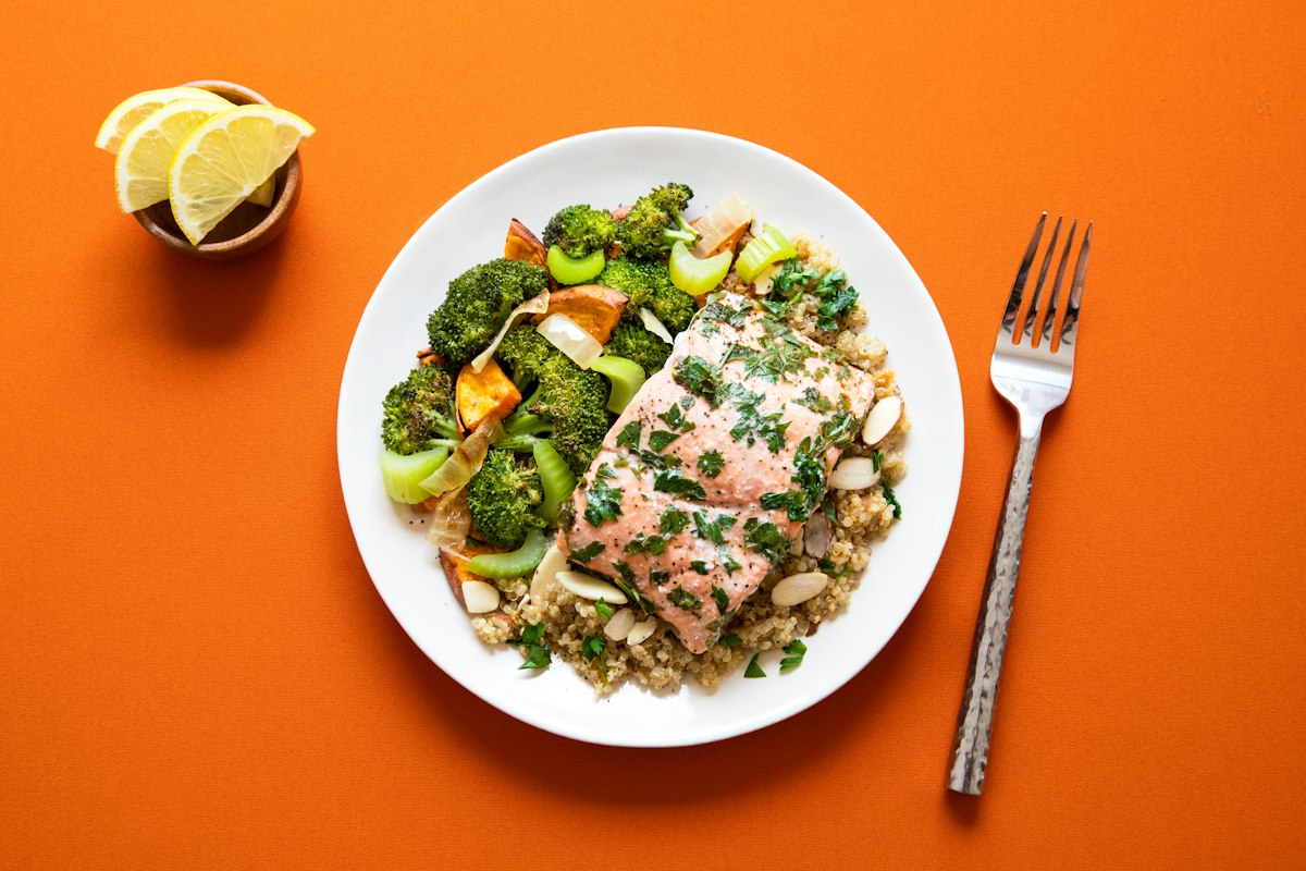 A salmon fillet on quinoa beside roasted broccoli and sweet potato, on an orange table