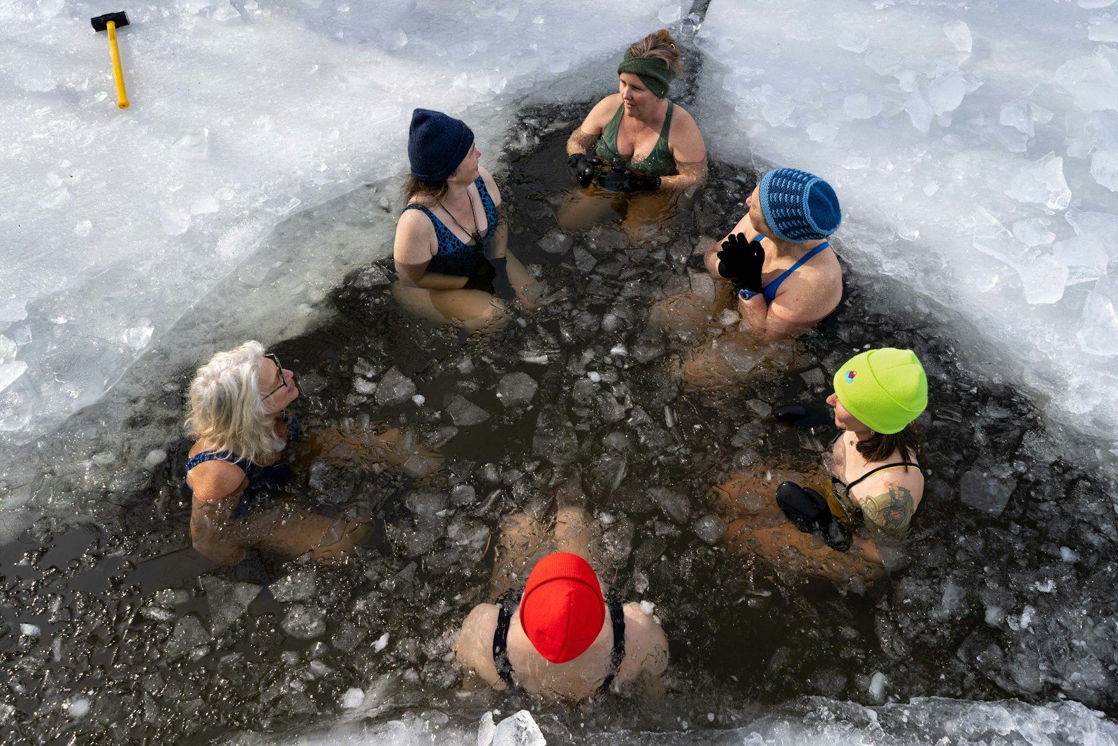 Six swimmers in woollen hats sitting in an ice hole, seen from above