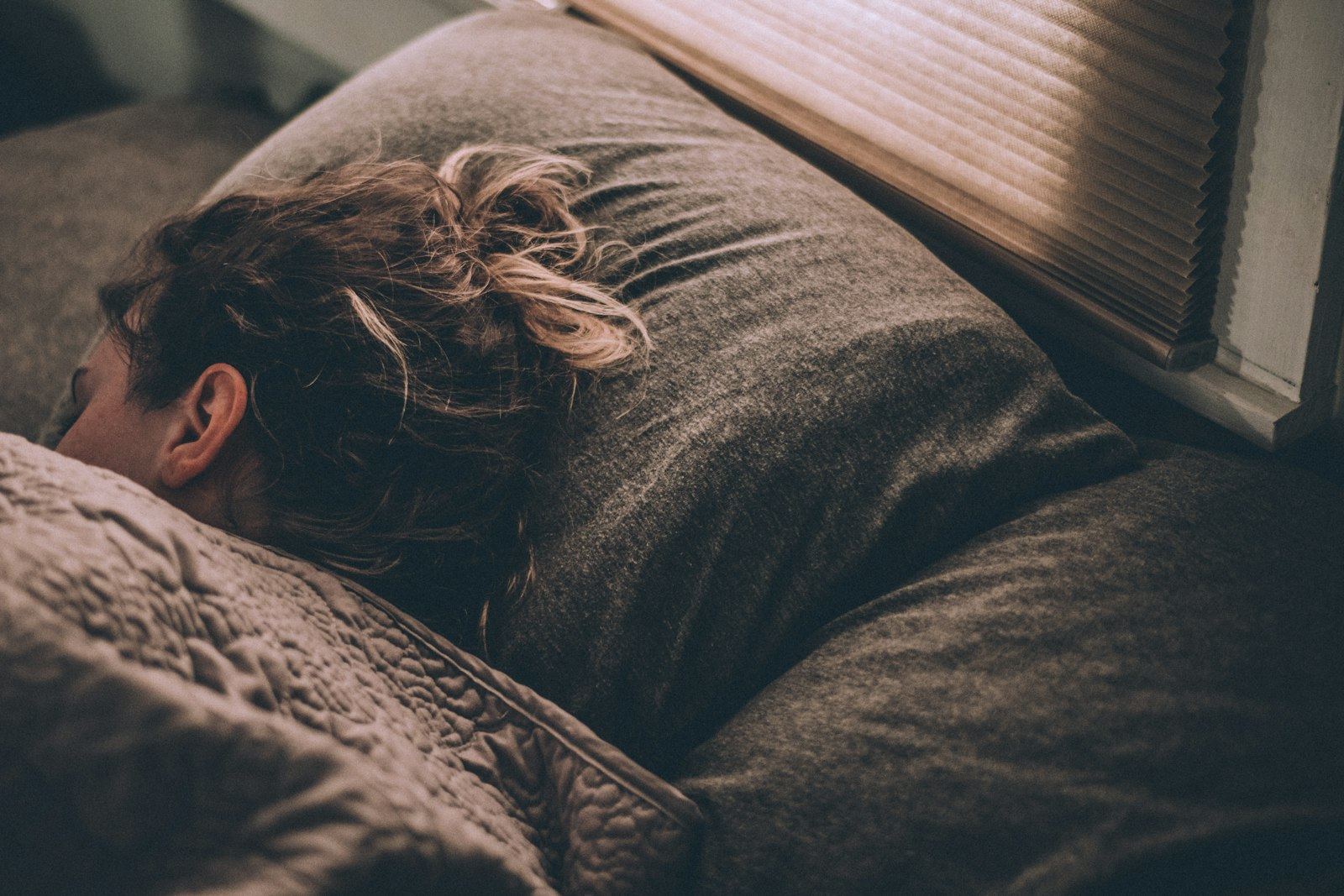 A person asleep face down in brown bedding, in low window light